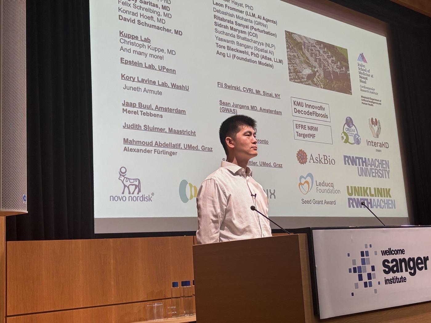 A speaker at the lectern of the Wellcome Sanger Institute in front of a slide listing collaborators and funders.